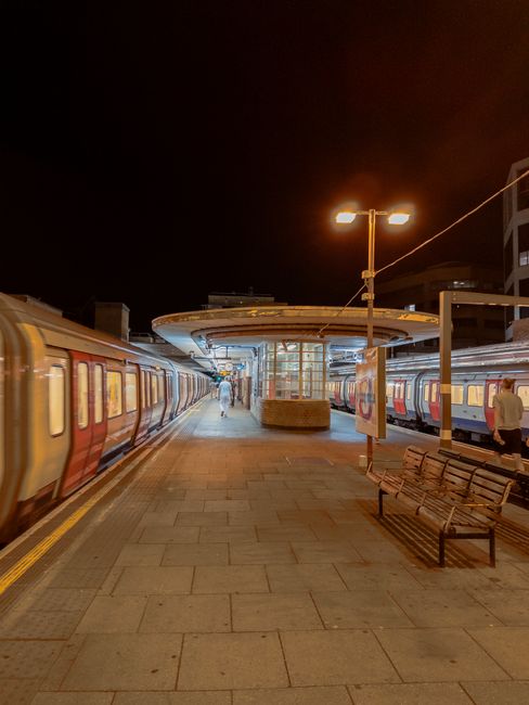 A spacious underground subway corridor with cream-colored tiled walls and a tiled floor, illuminated by evenly spaced ceiling lights. The tunnel features curved architectural elements on both sides an