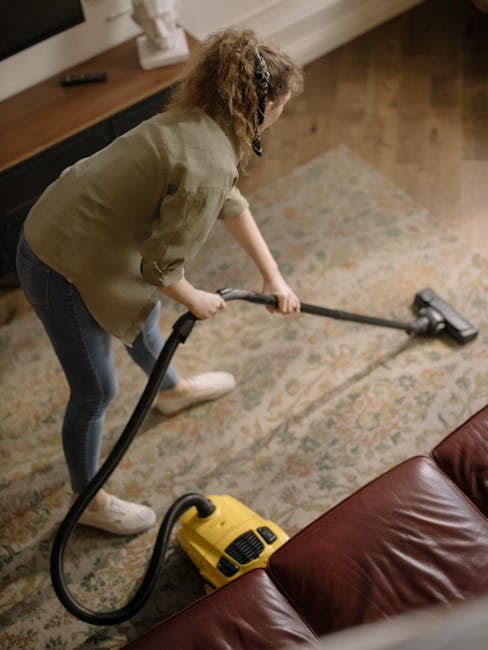 A professional cleaner from Creekmouth Carpet Cleaners is using a handheld electric upholstery steamer to deep clean the dark grey fabric sofa in a living room. The cleaning process involves careful a