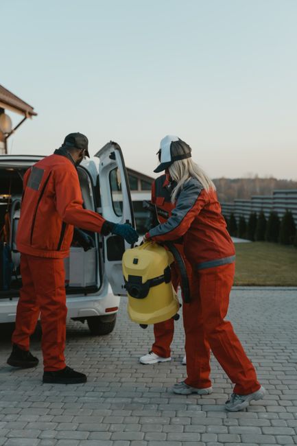 Two commercial cleaning professionals wearing orange high-visibility uniforms and protective gloves are preparing cleaning equipment from the back of a white van outdoors, near a well-maintained lawn